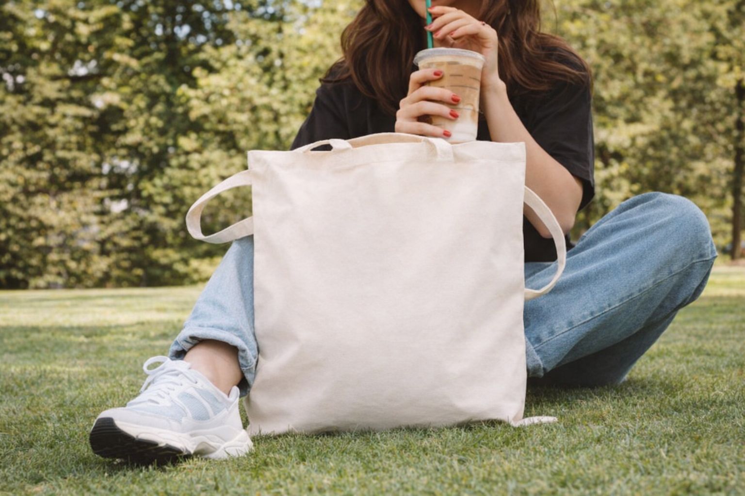 En student med en beige tygpåse och iskaffe i en grönskande park.